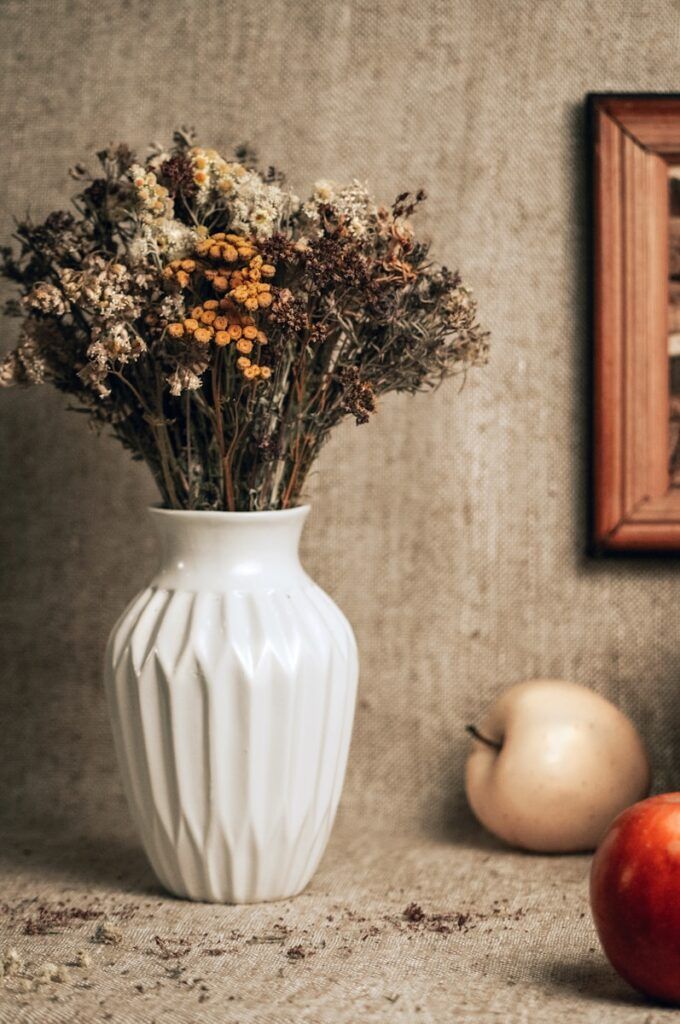 a white vase filled with dried flowers next to an apple