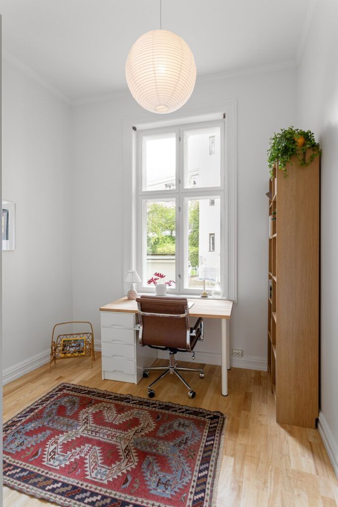 A room with a rug, a desk, white walls, wood flooring, and a window